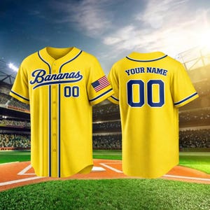 Puede incluir: Camiseta de béisbol amarilla con ribetes azules y la palabra "Bananas" en cursiva. La camiseta tiene el número "00" y un parche de la bandera estadounidense. La parte trasera de la camiseta tiene el texto "YOUR NAME" encima del número "00".