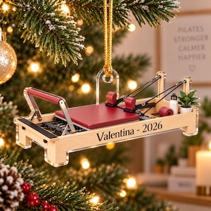 Könnte beinhalten: Ein Miniatur-Pilates-Reformer-Ornament mit einem hellen Holzrahmen, einem dunkelroten Schlitten, schwarzen Riemen und roten Schulterstützen. Die Seite des Rahmens ist mit dem Text 'Valentina - 2026' graviert. Es hängt an einer goldenen Kordel.
