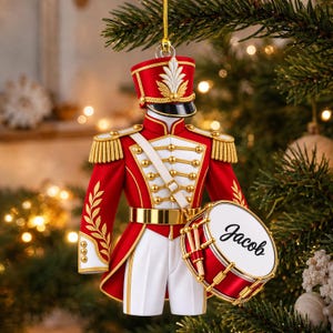 Può includere: Un ornamento a forma di uniforme da schiaccianoci rosso e bianco con spalline, bottoni e cintura dorati. L'uniforme include un cappello abbinato con un pennacchio bianco e un tamburo laterale con il nome Jacob scritto in nero.
