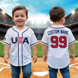 May include: A white baseball jersey with blue trim and the word "USA" in red and blue. The jersey also features the American flag on the sleeve. The back of the jersey has the text "CUSTOM NAME" and the number "99" in red.