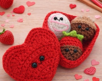 May include: A red crocheted heart-shaped box with a smiling face, open to reveal a white crocheted egg, a brown acorn, and a red strawberry. The box and contents are handmade. Scattered around are small heart-shaped decorations and a fresh strawberry.