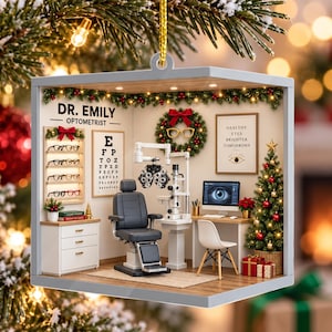 May include: A square Christmas ornament featuring a miniature optometry office. It includes a grey exam chair, a phoropter, a desk with a computer monitor displaying an eye, a wall-mounted eye chart, a shelf of glasses, and a decorated Christmas tree.