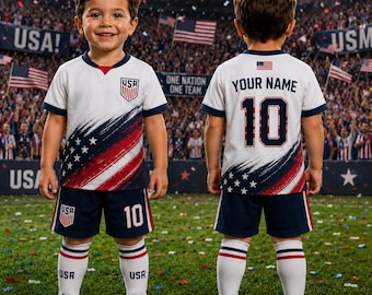 May include: A white soccer jersey and shorts set featuring red and blue brushstroke accents, white stars, and the USA crest. The back of the jersey displays the text YOUR NAME and the number 10. White socks with red stripes and USA text complete the kit.