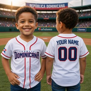 May include: Two children wearing white baseball jerseys with "República Dominicana" printed on the front. The jerseys have red and blue accents. The back of one jersey says "YOUR NAME" and "00". The background shows a baseball field and a banner that reads "República Dominicana".