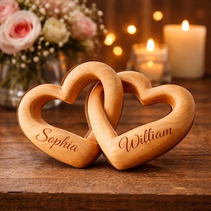 May include: Two wooden heart-shaped decorations interlocked, with the names "Sophia" and "William" engraved on each. The hearts are light brown and sit on a wooden surface. Soft lighting and flowers are in the background.