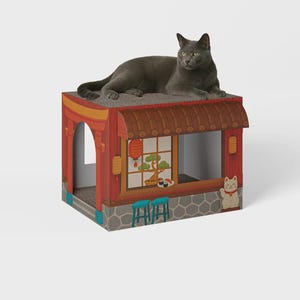 Op de afbeelding: Een grijze kat ligt op een kartonnen kattenhuis dat is vormgegeven als een Japans restaurant. De rood-bruine structuur heeft een raam met een bonsai-boom en sushi, en een gelukskattenfiguur.