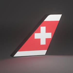 Pode incluir: Um estabilizador vertical de uma aeronave com fundo vermelho e uma cruz branca centrada na superfície. As bordas superior e inferior do estabilizador são finalizadas com uma faixa horizontal branca, criando um design geométrico distinto.