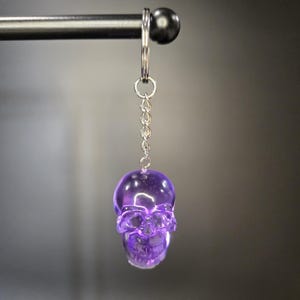 May include: A purple, translucent skull keychain hanging from a silver chain and ring. The skull has detailed facial features and is suspended against a blurred gray background. The keychain is attached to a black metal bar.