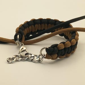 May include: A braided bracelet in black and brown, with a silver-colored chain clasp. The bracelet is made of woven cord, with a repeating pattern of black and brown sections. The clasp is a chain with a lobster claw closure.