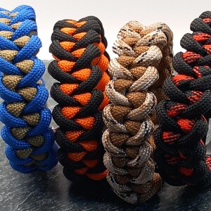 May include: Close-up of five braided paracord bracelets in various colour combinations. The bracelets feature blue and tan, black and orange, camouflage, and black with red accents. The braided design is visible.