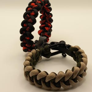 May include: Two paracord bracelets. One is black and red, the other is camouflage green and tan. Both bracelets have black clasps. The bracelets are made of woven nylon cord.