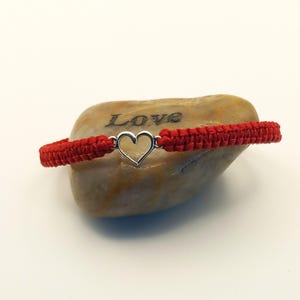 May include: Red macrame bracelet with a silver heart charm. The bracelet is displayed on a light brown stone with the word "Love" engraved on it. The bracelet is a handmade accessory.