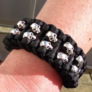 May include: Black paracord bracelet with silver skull beads. The bracelet is made of woven black cord and has multiple rows of skull-shaped beads evenly spaced across the top. The skulls have a detailed design.