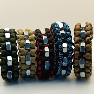 May include: Five paracord bracelets in different colours: olive green, tan, black with red accents, blue with black accents, and brown with green accents. Each bracelet has silver-coloured metal nuts. The bracelets are shown against a white background.