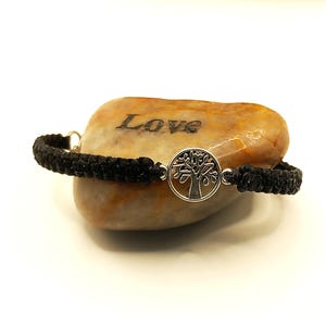 May include: A black braided bracelet featuring a silver tree of life pendant. The bracelet is presented on a light brown stone, engraved with the word "Love". This bracelet is a simple, elegant piece of jewellery.