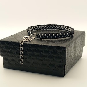 May include: A black and white braided collar with a silver-tone chain and clasp. The collar is resting on a black, textured box. The collar is designed with a repeating pattern of black and white strands.