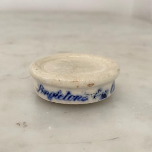 May include: A small, round, off-white ceramic lid with a slightly scalloped edge. The word "Pingietons" is written in blue script around the side. The lid appears to be vintage or antique.