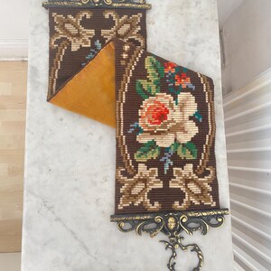 May include: A vintage tapestry wall hanging with a floral design. The tapestry has a brown background with a detailed pattern and a large flower with red and white petals. It is framed with ornate brass hardware.