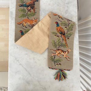 May include: A vintage tapestry with embroidered wildlife scenes. The tapestry shows a pheasant, fox, and dog, set against a neutral backdrop. It has a braided handle and a tassel with colourful threads.