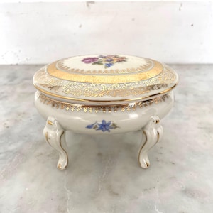 May include: A small, white porcelain trinket box with a floral design on the lid. The lid is trimmed with gold filigree and a gold band. The box has three curved feet and a blue floral accent on the base. A decorative piece for storing jewelry or small items.