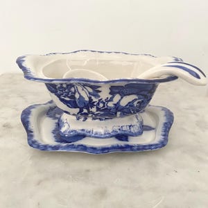 May include: A white and blue ceramic serving bowl with a matching spoon, resting on a small, rectangular dish. The bowl and dish feature a blue floral pattern. The spoon rests on the rim of the bowl. The set is designed for serving.