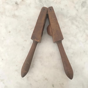 Könnte beinhalten: Ein Vintage-Nussknacker aus Holz mit Hebeldesign. Der Nussknacker ist aus dunkelbraunem Holz gefertigt und hat ein einfaches, funktionales Design. Die Griffe sind abgerundet und verjüngt, und die Backen sind zum Knacken von Nüssen ausgelegt. Der Nussknacker ist wahrscheinlich ein Antiquität.