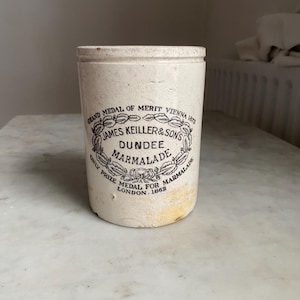 May include: An antique, off-white ceramic marmalade jar. The jar features a black and white circular emblem with text that reads "JAMES KEILLER & SON'S DUNDEE MARMALADE" and "GRAND MEDAL OF MERIT VIENNA 1873".