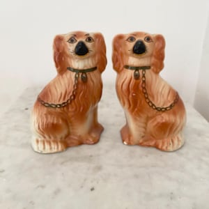 May include: Two ceramic dog figurines, each with a light brown coat and black nose. The dogs are seated, with detailed fur texture and a decorative chain collar. The figurines are on a marble surface.