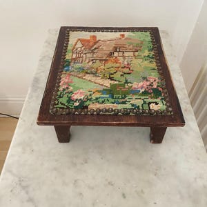 May include: A small, wooden footstool with a needlepoint top. The needlepoint shows a detailed landscape scene with a house, trees, and flowers. The frame is dark brown, and the needlepoint is secured with small, round studs.