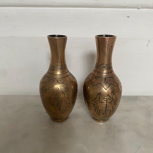 May include: A pair of antique bronze vases with a slender neck and a rounded body. Each vase has etched designs of birds and other Asian-inspired motifs. The vases are displayed on a marble surface.