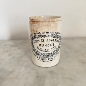 May include: A cylindrical off-white ceramic marmalade jar with a visible crackled glaze finish. The front features black printed text reading James Keiller & Sons Dundee Marmalade, along with references to prize medals from Vienna and London 1862.