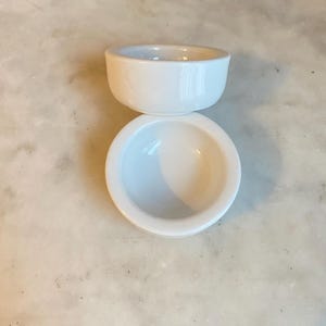 May include: Two small, white ceramic bowls are stacked. The top bowl is upright, the bottom one inverted. The bowls have a simple, rounded design and a glossy finish. They are likely for serving dips or small portions. The bowls are made of ceramic.