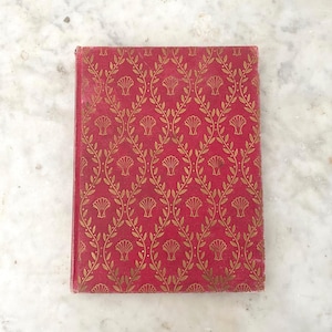 May include: A vintage book with a red cover and a gold leaf and shell pattern. The rectangular cover shows signs of wear. The design is symmetrical and ornate, giving the book a classic aesthetic. The book's cover is red with a gold pattern.