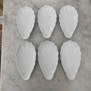 May include: Six white, shell-shaped serving dishes on a marble surface. Each dish has a detailed, ribbed design, resembling a scallop shell. The dishes are made of porcelain or a similar material.