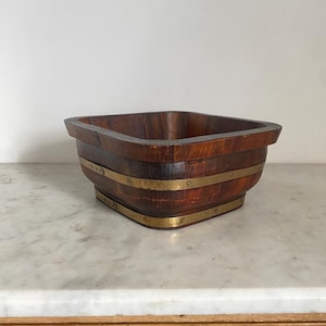 May include: A square wooden bowl with a dark brown finish, featuring two horizontal brass bands secured with small metal rivets. The bowl has a wide, flat rim and a tapered base, showcasing a natural wood grain texture throughout the exterior.