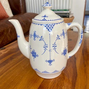 May include: A white porcelain teapot with a lid, decorated with blue floral patterns and accents. The teapot has a curved spout and a handle, and it sits on a wooden surface. The design is classic and elegant.