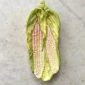 May include: A ceramic corn-on-the-cob shaped dish. The dish features two ears of corn with white kernels and red silk, wrapped in green husks. The dish is painted with a glossy finish.