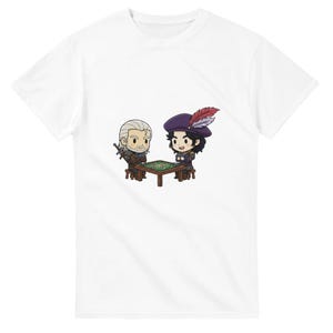 May include: White t-shirt featuring a cartoon illustration of two characters seated at a table. One character has white hair and a sword, the other wears a purple hat with a feather. They are playing a game on a green table.