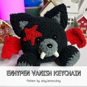 Pode incluir: Um chaveiro de pelúcia preto feito à mão em forma de criatura semelhante a um lobo, com asas vermelhas e uma estrela vermelha. O chaveiro tem detalhes cinzentos e presas brancas. O texto "ENHYPEN VANISH KEYCHAIN" é exibido abaixo.