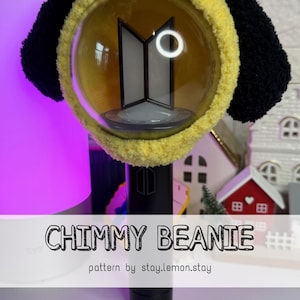 Puede incluir: Un palo de luz negro con un gorro Chimmy amarillo. El gorro tiene detalles negros en forma de orejas y una textura esponjosa. El palo de luz tiene un globo transparente con un logotipo y el texto "CHIMMY BEANIE" se muestra.