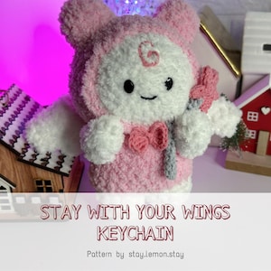 May include: A handmade pink and white plush keychain shaped like a character with a hood, wings, and a bow. The keychain has the text "STAY WITH YOUR WINGS KEYCHAIN".