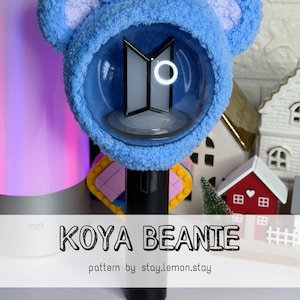 May include: A blue Koya Beanie, a plush bear-eared cover for a light stick. The beanie has a fluffy texture and a clear dome to showcase the light stick's design. Text on the image reads "KOYA BEANIE" and "pattern by stay.lemon.stay."