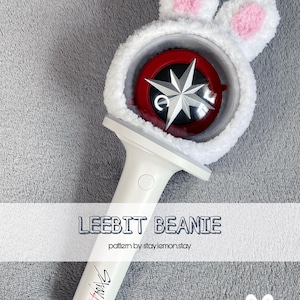 May include: White and red light stick with a bunny ear beanie. The beanie has pink inner ears and a fluffy texture. The light stick handle is white with the text "Stray Kids". The text "LEEBIT BEANIE" is also visible.