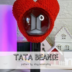 May include: A red, heart-shaped beanie covers a clear, spherical object with a white logo. The beanie has a fluffy texture and the text "TATA BEANIE" is displayed on the image, along with "pattern by stay.lemon.stay".
