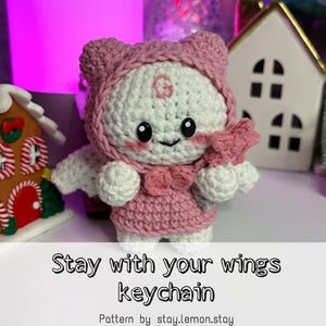 Pode incluir: Um chaveiro de crochê feito à mão com um personagem branco e rosa com laço, asas e capuz. O chaveiro tem olhos pretos e um rosto sorridente. O texto "Stay with your wings keychain" é exibido abaixo.