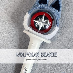 May include: A light blue and navy blue crocheted Wolfchan beanie, designed to fit over a light stick. The beanie has wolf ears and a white trim. The light stick has a red and silver design and the text "Stray Kids".