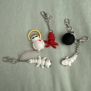May include: A collection of handmade keychains featuring various designs. Includes a white robot with a gold ring, a red octopus, a black ball, a white segmented creature, and a white fish with red accents. All keychains have silver-colored clasps.