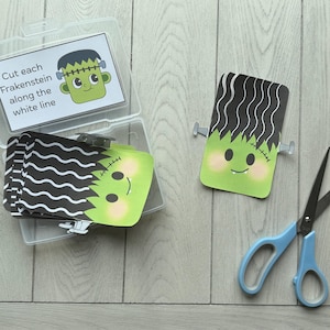 May include: A clear plastic container holds a stack of Frankenstein-themed cards with black wavy hair and green faces. One card is separate, and a pair of blue scissors rests nearby. Text on a card reads, "Cut each Frankenstein along the white line."