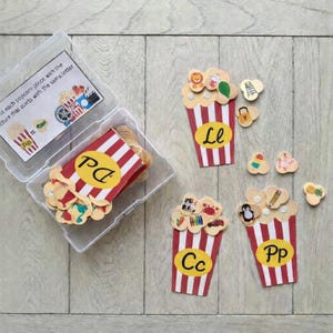 Op de afbeelding: Educatief spel met popcorn-thema. De set bevat rood-wit gestreepte popcornbakjes met letters en kleine plaatjes. Het spel is ontworpen om de plaatjes aan de letters te koppelen. De tekst op de doos luidt: "Match elk popcornstuk met de afbeelding die met dezelfde letter begint."