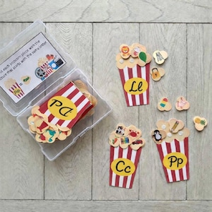 Op de afbeelding: Educatief spel met popcorn-thema. De set bevat rood-wit gestreepte popcornbakjes met letters en kleine plaatjes. Het spel is ontworpen om de plaatjes aan de letters te koppelen. De tekst op de doos luidt: "Match elk popcornstuk met de afbeelding die met dezelfde letter begint."